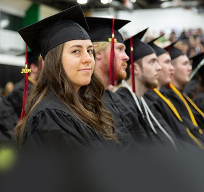 Row of new Gateway graduates sitting at commencement in caps and gowns, the closest smiling.